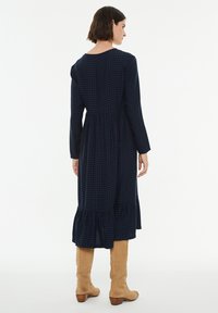 Vestido a cuadros en azul marino y verde oscuro con mangas largas, un cuerpo ajustado y un dobladillo acampanado, combinado con botas de caña alta en color beige.