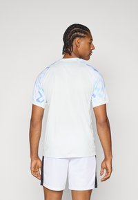 T-shirt de sport gris clair avec un motif géométrique bleu sur les épaules, manches courtes, col rond, associé à un short blanc.