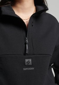 Pullover in pile nero con zip e colletto alto, dotato di tasca frontale, tessuto testurizzato e una patch intrecciata con il logo "Superdry".