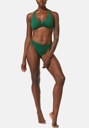 Femme portant un bikini deux pièces vert avec des détails anneaux, debout avec une jambe légèrement levée sur un fond blanc uni.