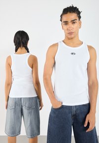 White ribbed tank top with round neckline, featuring a small black logo. Paired with loose, knee-length denim shorts in light and dark washes.