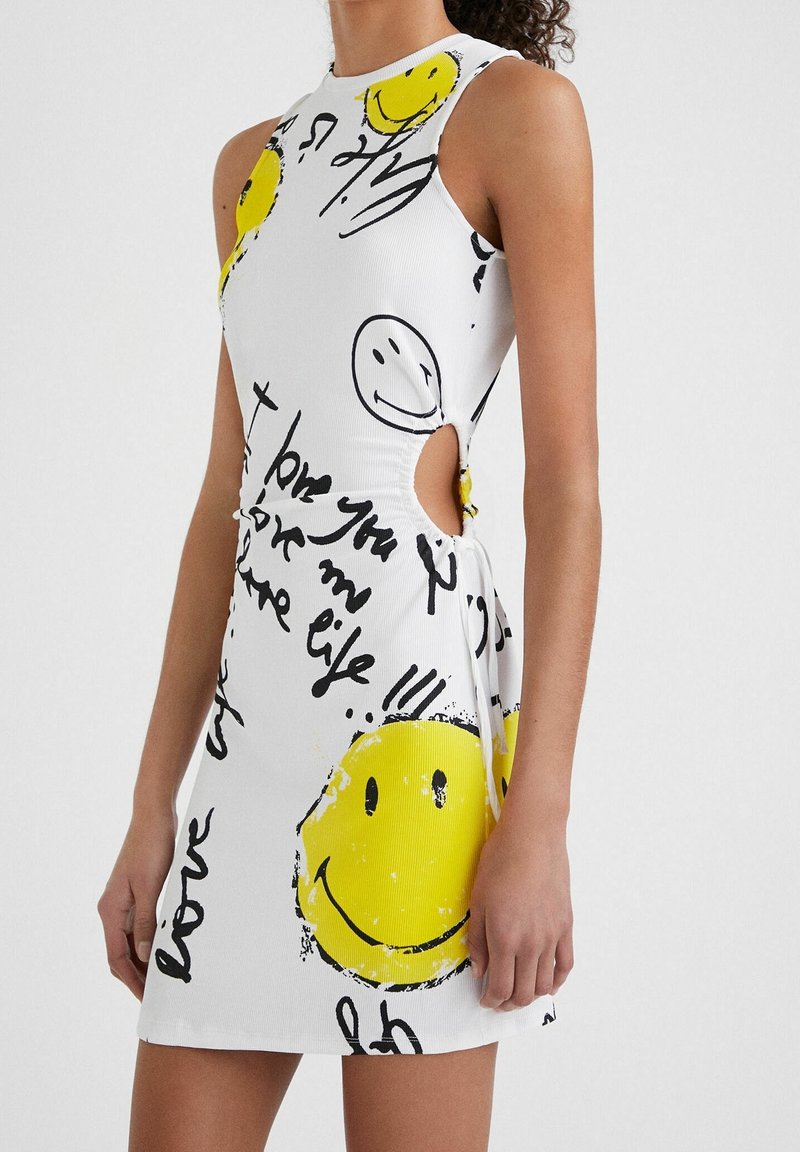 Robe blanche sans manches avec de grands visages souriants jaunes, texte manuscrit noir et détail découpé sur le côté, portée par une personne debout.