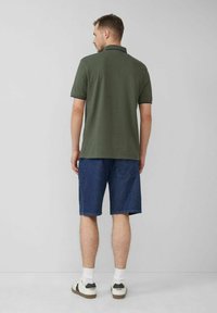 Dunkelgrünes Polohemd mit kurzen Ärmeln, kombiniert mit Jeansshorts. Legere Schuhe und weiße Socken runden den Look ab. Einfacher Hintergrund.