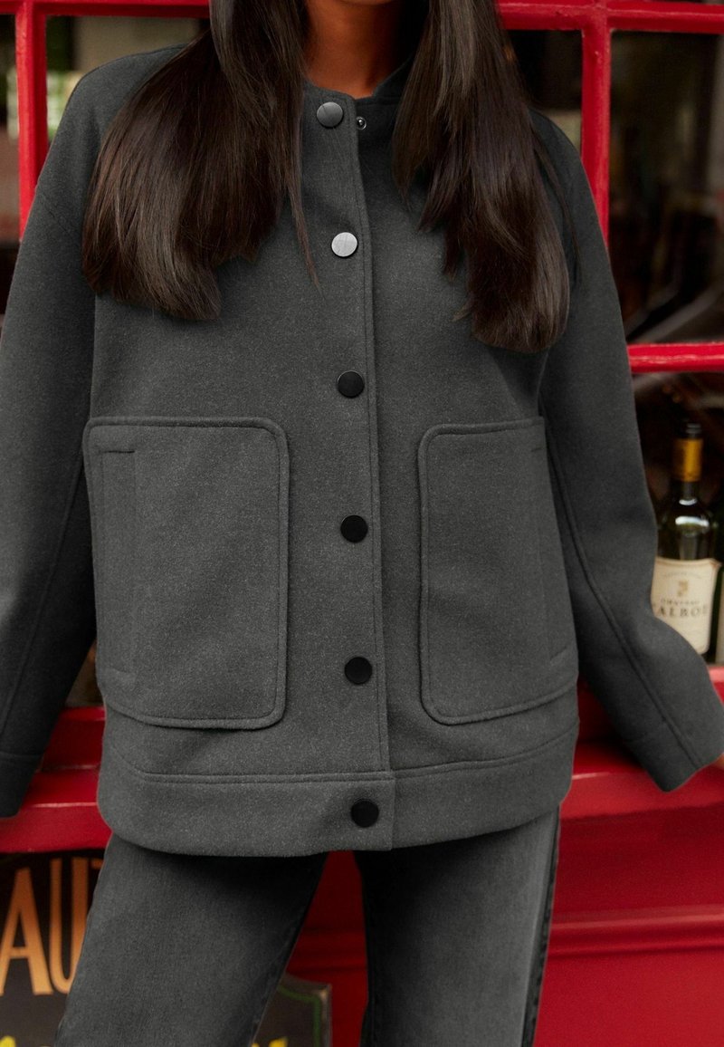 Veste en mélange de laine grise avec un col arrondi, une fermeture à boutons noirs sur le devant et deux grandes poches plaquées.