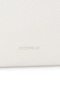 Bílá kožená peněženka s texturovaným povrchem, se zlatým embosovaným logem "Coccinelle". Kompaktní design s čistými liniemi.