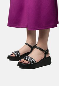 Czarne sandały na platformie z zaokrąglonym czubkiem, skórzanymi paskami i zapięciem na klamrę. Posiadają białą taśmę z logo na górze.