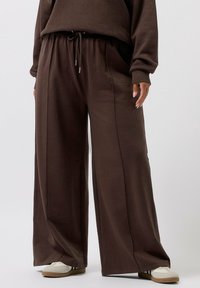 Braune, weite Hosen mit elastischem Bund, ausgestattet mit Kordelzug und seitlichen Taschen. Weicher Stoff mit glatter Textur.