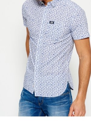 Homme portant une chemise blanche à manches courtes avec un motif floral bleu et un jean bleu, debout devant un fond blanc uni.