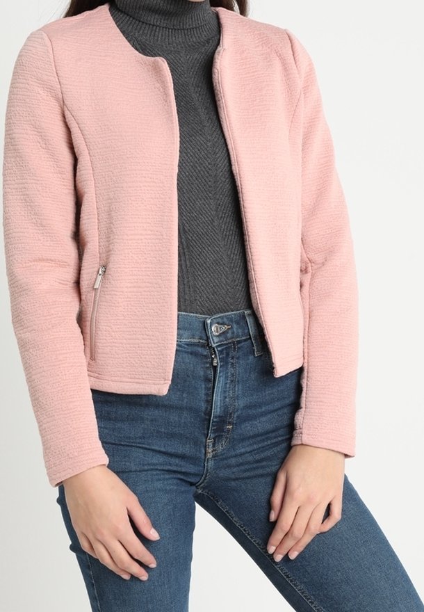 Veste rose pâle texturée avec un col rond, une fermeture éclair à l'avant et deux poches latérales. Portée sur un col roulé gris foncé. Coupe ajustée.