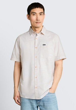 Homme portant une chemise beige clair à manches courtes avec boutons sur le devant et une poche avant, et un jean bleu clair, debout devant un fond uni.