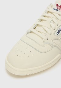 Valge nahast toss, millel on auguline varvas, hallid aktsendid ja valged paelad. Sisaldab Adidas logo sinises värvuses küljel ja keelel.