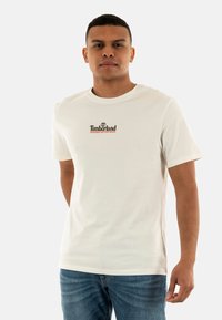 Camiseta de algodón blanca con mangas cortas y escote redondo. Logo destacado en texto marrón y naranja en el pecho. Lleva puesta unos jeans azules.