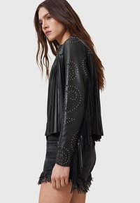 Juoda odinė striukė su kutais per rankoves, dekoruota kniedėmis ir trumpintas modelis, derinamas su nudėvėtais džinsiniais šortais.