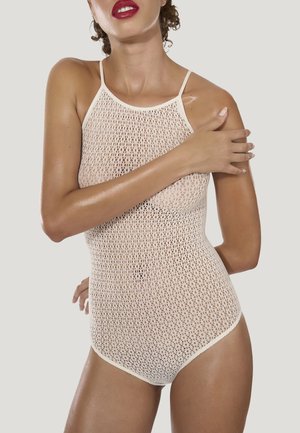 Femme en body beige sans manches en maille avec bretelles spaghetti, posant avec la main sur l'épaule opposée devant un fond uni.