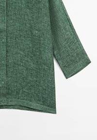 Chemise à manches courtes avec boutonnière, en tissu texturé vert foncé, présentant un ourlet droit et un léger éclat. Les boutons sont assortis à la couleur du tissu.