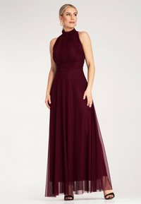 Burgunderfarbene Maxikleid mit Halsband, körperbetontem Oberteil und fließendem Rock, aus leichtem Stoff gefertigt. Schwarze Riemchen-High Heels runden den Look ab.
