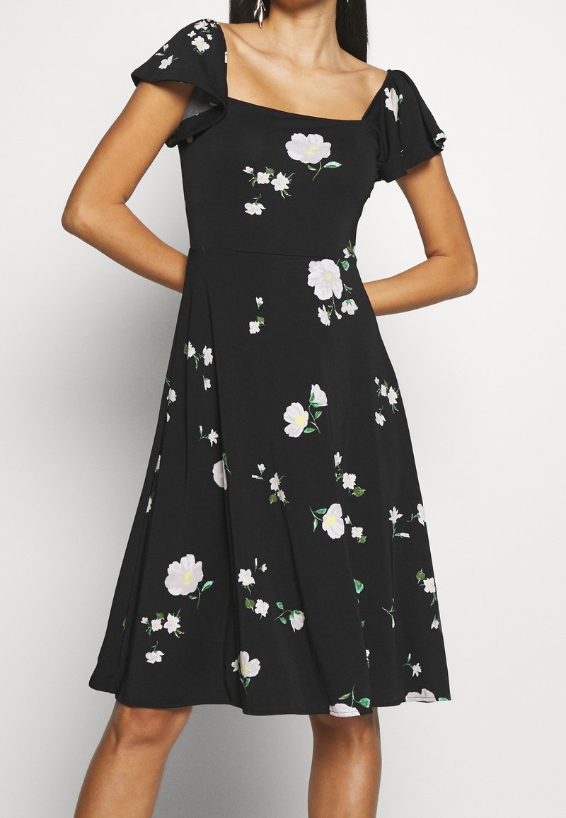 Schwarzes Kleid mit quadratischem Ausschnitt, kurzen ausgestellten Ärmeln und Blumenmustern in Weiß und Grün. Der ausgestellte Rock reicht bis zur Mitte der Wade.