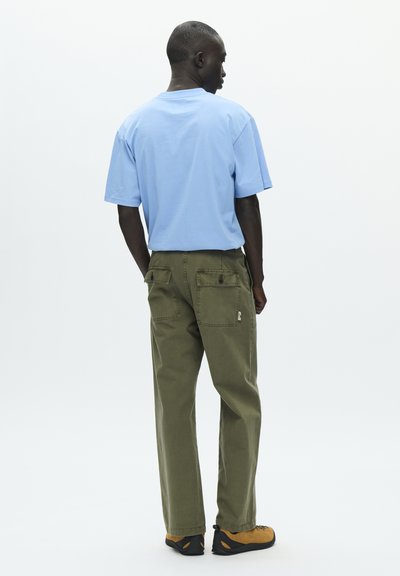T-shirt en coton bleu clair, coupe décontractée. Pantalon utilitaire vert olive avec plusieurs poches et quincaillerie noire. Chaussures marron avec semelles noires.