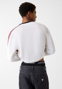 Biała siateczkowa koszula z długim rękawem, czarnym kołnierzem i czerwonym akcentem na ramieniu, w połączeniu z ciemnymi jeansami z pasem w talii z logo.