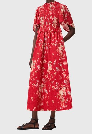 Vrouw draagt een rode midi-jurk met bloemenprint, pofmouwen en een geplooide taille, gecombineerd met zwarte platte sandalen tegen een effen achtergrond.