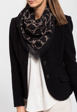 Femme portant un blazer noir sur un chemisier blanc avec une écharpe Calvin Klein noire et beige à motifs autour du cou.