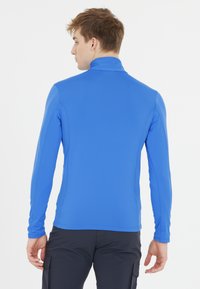 Langærmet blå træningsskjorte med høj krave og glat tekstur, kombineret med mørke bukser. Har tætsiddende design og flade sømme.