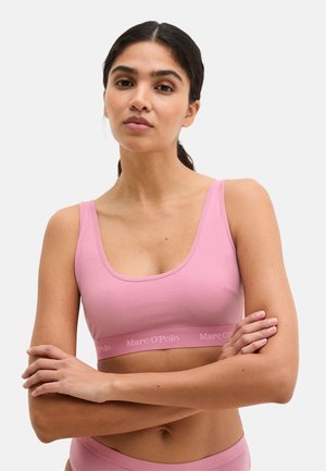Frau mit dunklen Haaren zu einem niedrigen Pferdeschwanz gebunden, trägt einen rosa Sport-BH von Marc O'Polo und passende Unterwäsche, mit verschränkten Armen und neutralem Gesichtsausdruck.