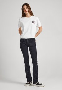 Bela majica s kratkimi rokavi z črnim logotipom "Pepe Jeans", v kombinaciji z temno modrimi tesnimi jeans hlačami in črnimi supergami z belimi podplati.
