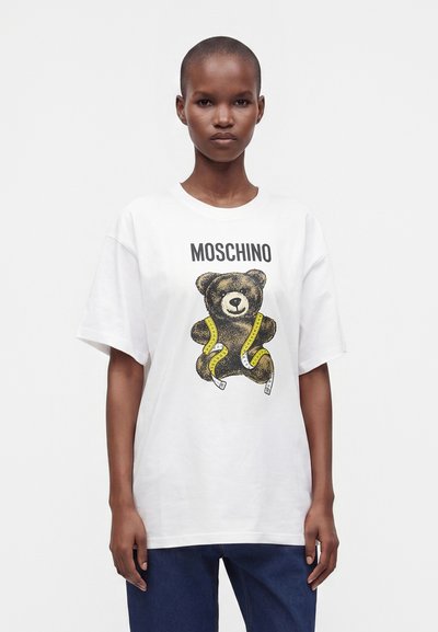 Άτομο με κοντά μαλλιά που φοράει ένα λευκό t-shirt Moschino με μια αρκουδάκι τυλιγμένη με κίτρινη μεζούρα.