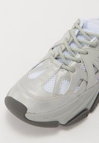 Zapatilla deportiva gris claro con superposiciones de malla y sintético, con un cuello acolchado, cordones grises y una suela de goma negra texturizada.