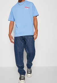 Ljusblå bomullströja med trycket "on vacation CLUB", kombinerad med löst sittande mörkblå jeans och svarta och vita sneakers.