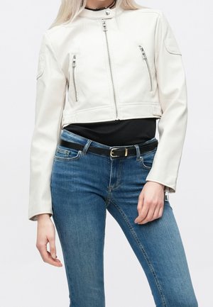 Femme portant une veste courte blanche à fermeture éclair devant, un haut noir, un jean bleu et une ceinture noire, debout devant un fond uni.