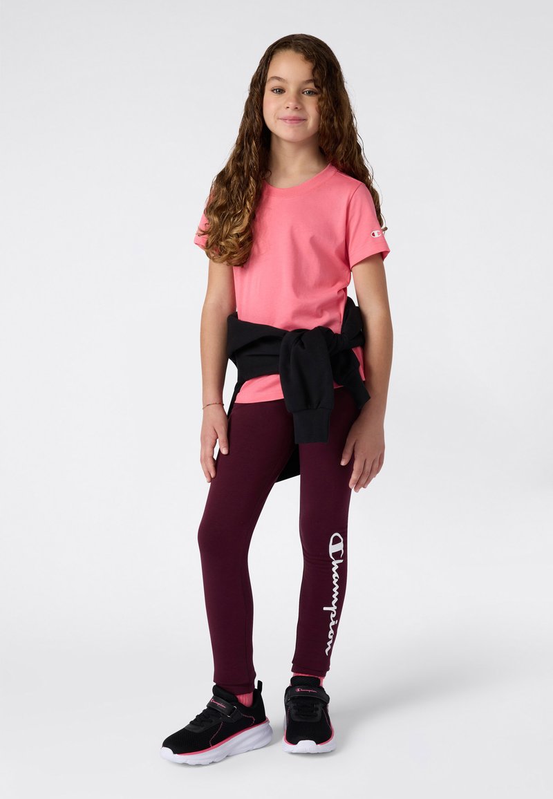 Różowa koszulka z krótkim rękawem, czarne legginsy z białym logo i czarne buty sportowe z różowymi akcentami. Modelka stoi przodem.