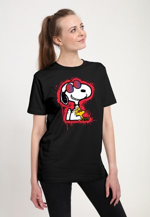 Ung kvinde med ponytail iført sort t-shirt med Snoopy og Woodstock tegneserie i solbriller på rød baggrund.