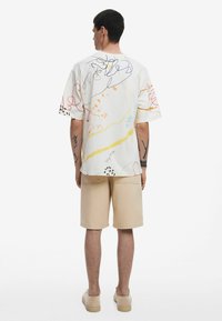 Maglietta a maniche corte con collo tondo in tessuto bianco con design astratti multicolori; abbinata a pantaloni corti beige e scarpe casual.