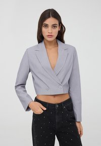 Giacca grigia tagliata con chiusura a doppio bottone, rever a punta e maniche lunghe, abbinata a jeans neri con decorazioni bianche.