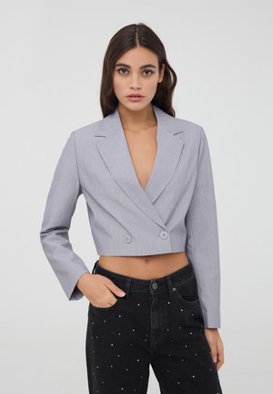 Giacca grigia tagliata con chiusura a doppio bottone, rever a punta e maniche lunghe, abbinata a jeans neri con decorazioni bianche.