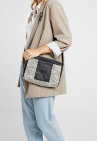 Donna che indossa un blazer beige a righe e jeans chiari, mentre tiene una borsa a tracolla nera e bianca con tasca frontale.