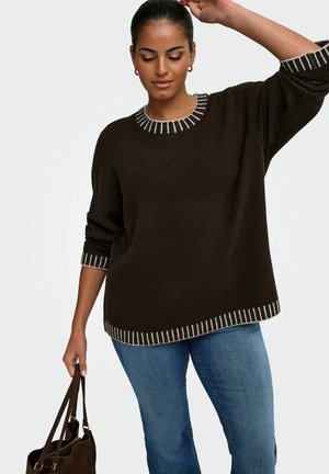 Donna che indossa un maglione marrone scuro con bordi bianchi e jeans blu, che tiene una borsa marrone, posando con un braccio sollevato sopra la testa.