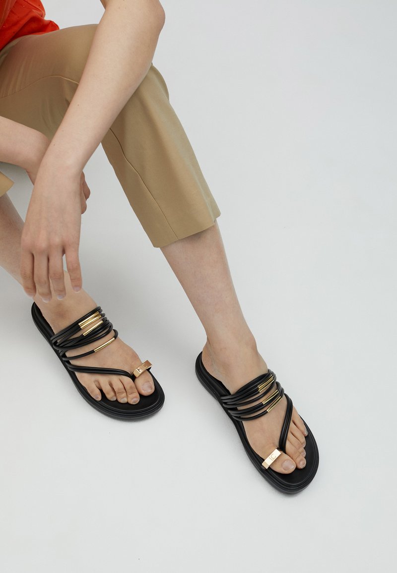 Mustat flip-flop-sandaalit, joissa on useita toisiinsa kietoutuneita hihnoja ja kultaisia yksityiskohtia. Pinta on teksturoitu takaamaan pitoa. Yhdistetty vaaleanruskeisiin tai lyhyisiin housuihin.