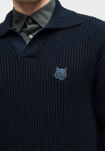 Homme portant une chemise boutonnée vert foncé sous un pull en maille bleu marine avec une petite tête de renard brodée sur la poitrine.