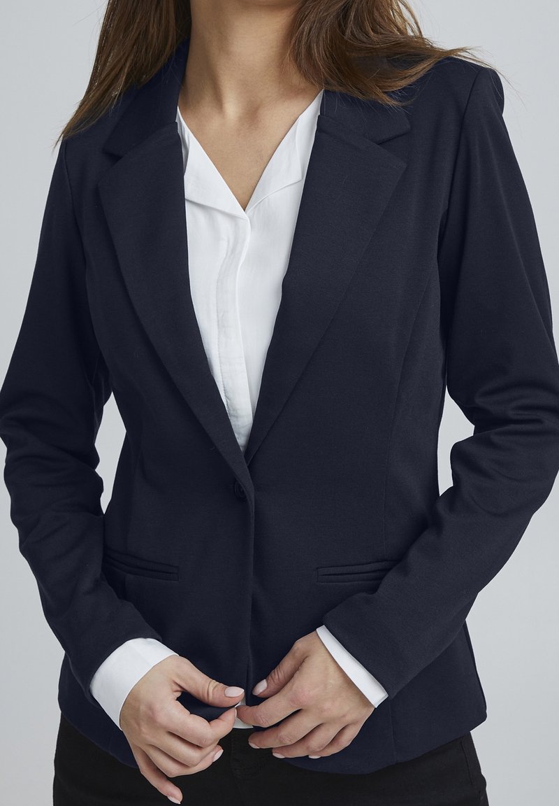 Femme portant un blazer bleu marine ajusté sur une chemise blanche à col, boutonnant le blazer avec les deux mains.