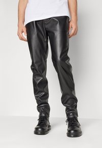 Man in zwarte faux leren broek en zwarte veterschoenen, staand tegen een effen lichte achtergrond.