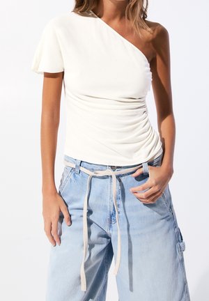 Woman wearing a white one-shoulder ruched top and light blue high-waisted jeans with a thin fabric belt tied in front.