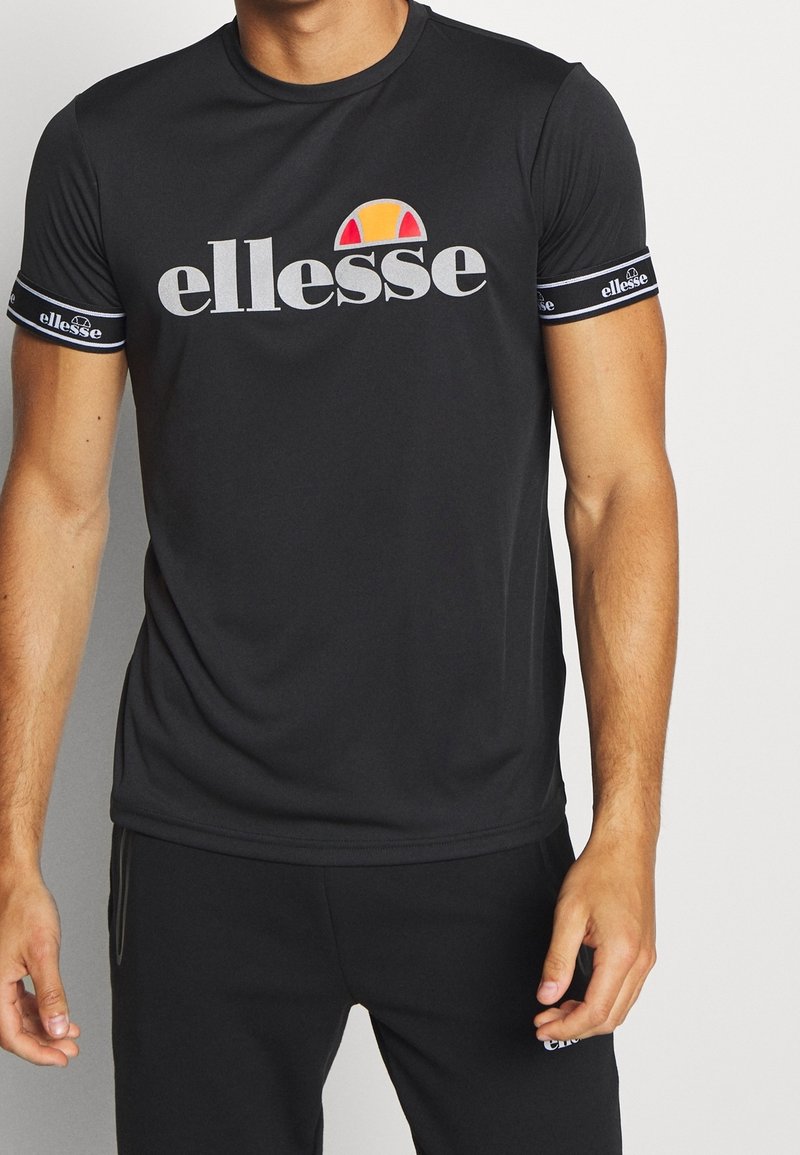 Černé tričko s krátkým rukávem s velkým bílým logem "ellesse" a barevným emblémem, s pruhovanými akcenty na rukávech.