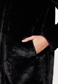 Manteau en fausse fourrure noire avec une texture douce et duveteuse. Présente une poche latérale et un design décontracté. Convient pour un look décontracté ou confortable.