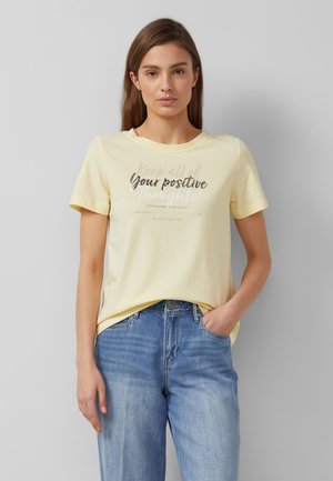 Frau mit glatten braunen Haaren, die ein hellgelbes T-Shirt mit dem Text "Deine positiven Gedanken" und blaue Jeans trägt, steht vor einer hellgrauen Wand.