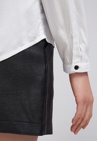 Chemise blanche légère à manches longues avec une finition texturée et un détail de bouton noir au poignet, associée à une jupe en cuir noire brillante.
