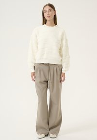 Cremfarbener Pullover mit wellenförmigem Muster, kombiniert mit beigen hoch taillierten, gefalteten Hosen und hellen Slippern.