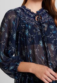 Námořnická květinová blůza s lehkou, průsvitnou látkou a modro-bílými květinovými vzory. Nařasený výstřih se zavazováním a nabírané rukávy.